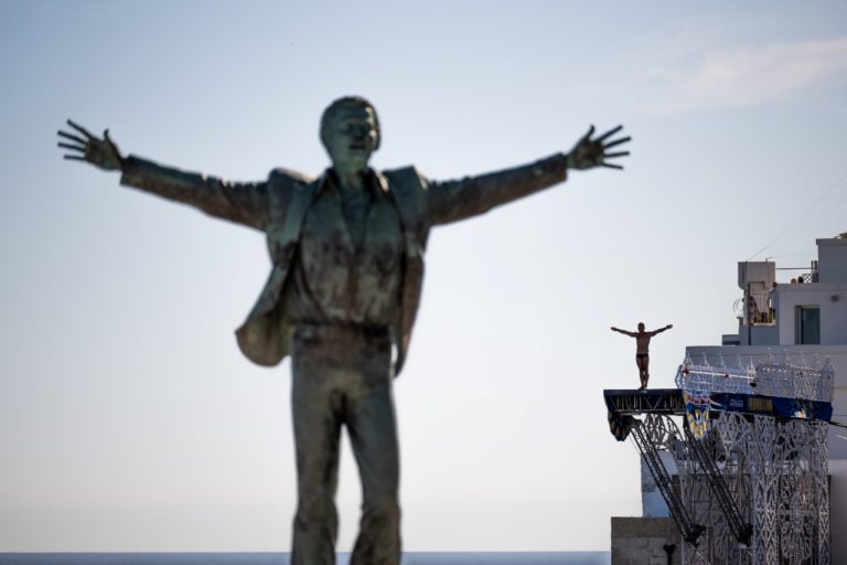 Alessandro De Rose of Italy prepares to dive from the 27 metre platform as Dario Costa (L) of Italy flies by the statue of Domenico Modugno during the Volare project prior to the seventh stop of the Red Bull Cliff Diving World Series in Polignano a Mare, Italy on September 14, 2022. // Romina Amato / Red Bull Content Pool // SI202209140484 // Usage for editorial use only //